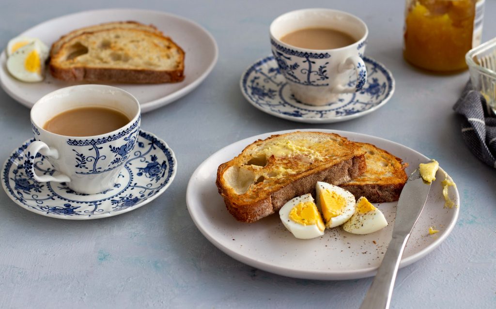 A breakfast setting with two cups of tea, along with a plate that has two slices of toast, three pieces of boiled eggs and a butter knife. 