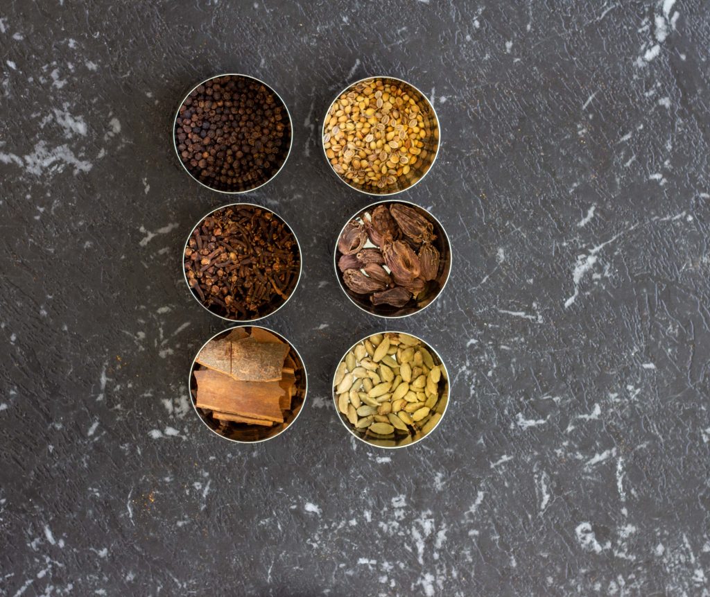 Whole spices that are used regularly in a Desi kitchen placed in small steel bowls - they include cinnamon sticks, cloves, cardamom, black peppercorn, black cardamom and fenugreek seeds.