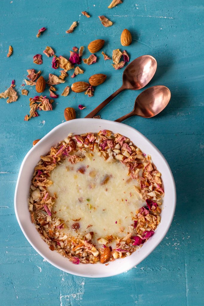 A bowl of chawal ki kheer (Pakistani rice pudding) at the bottom. The rice pudding is garnished with a circle of crushed almonds and dried rose petals. Above the bowl are two small bronze teaspoons and a scattering of edible rose petals and almonds. 