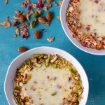 Two bowls of chawal ki kheer (Pakistani rice pudding). The bowl on the bottom left is garnished with crushed almonds and sliced pistachios. Above that, on the right corner is another bowl of the rice pudding, this one decorated with crushed almonds and dried rose petals. Next to the bowl on the left are scattered almonds, pistachios and dried rose petals.