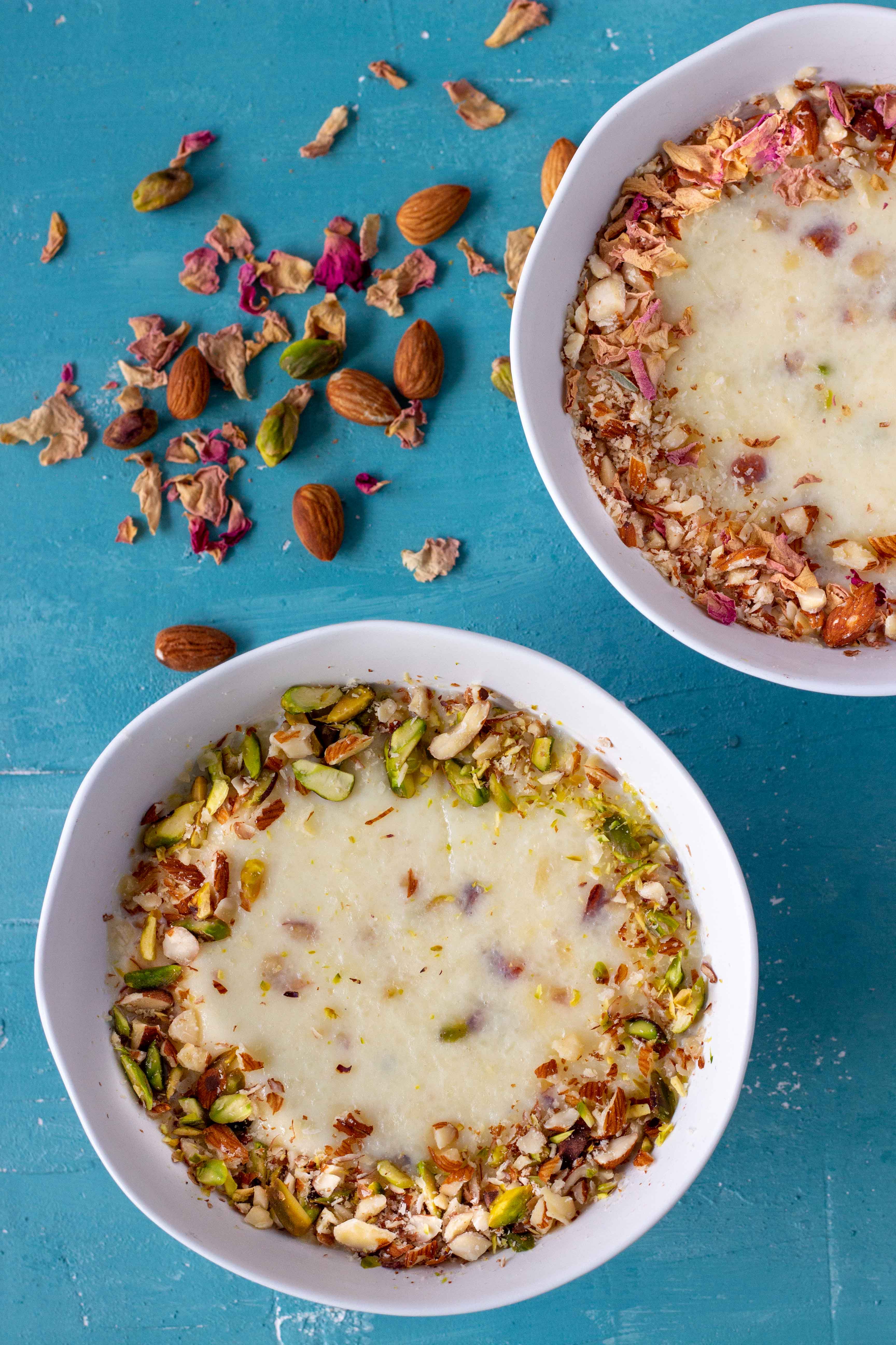 Two bowls of chawal ki kheer (Pakistani rice pudding). The bowl on the bottom left is garnished with crushed almonds and sliced pistachios. Above that, on the right corner is another bowl of the rice pudding, this one decorated with crushed almonds and dried rose petals. Next to the bowl on the left are scattered almonds, pistachios and dried rose petals.