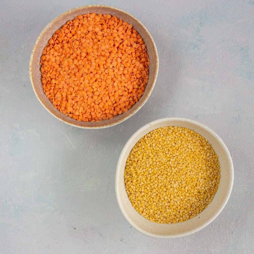 On the bottom right is a white bowl containing yellow lentils or moong daal. On the top left is a white bowl containing red lentils or masoor daal. 