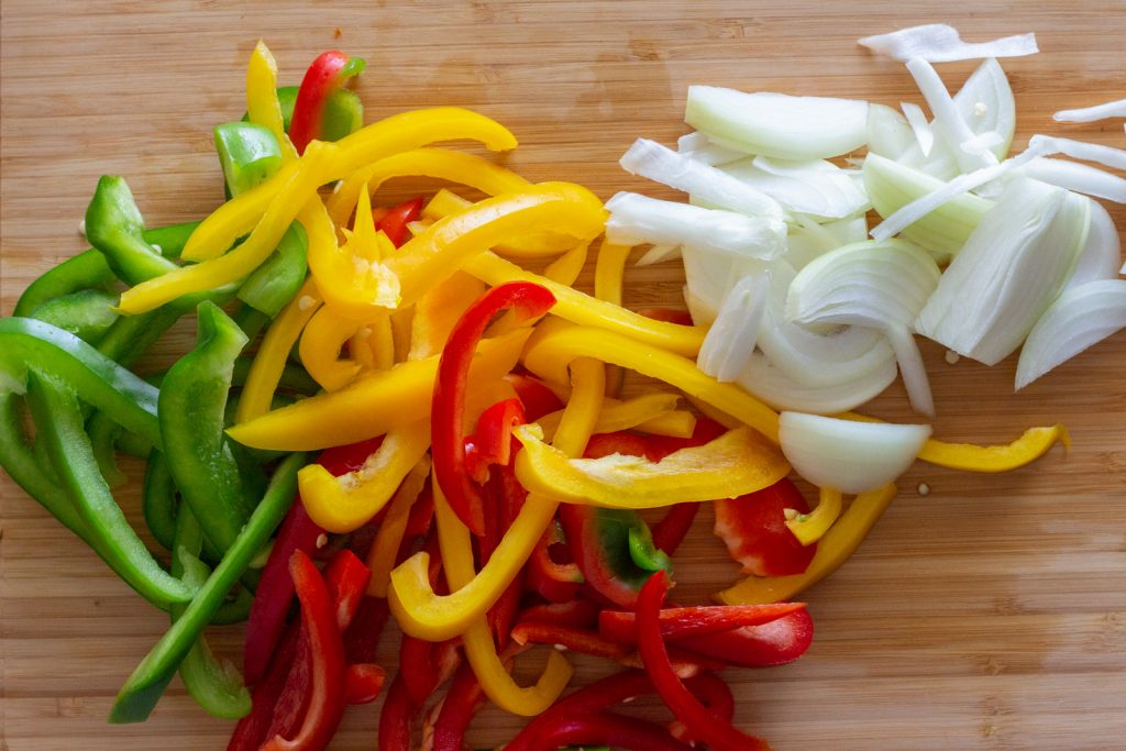 A chopping board with sliced bell peppers (red, yellow and green) along with sliced onions on it. 