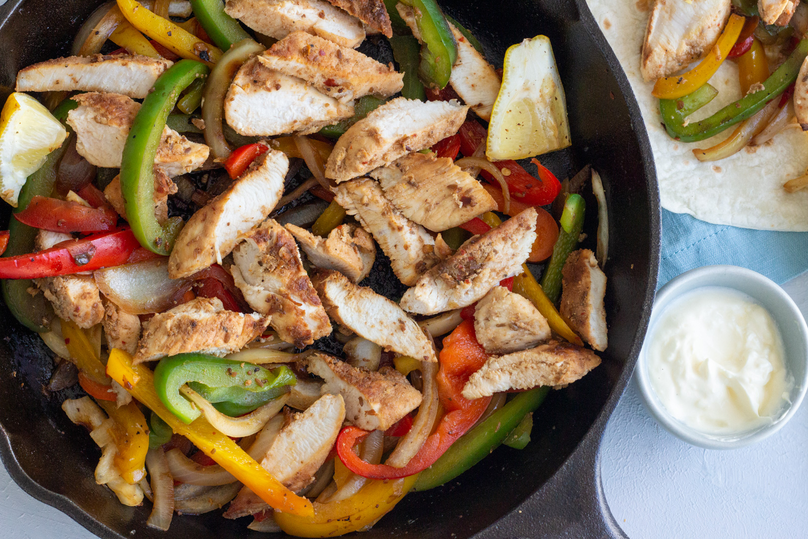 A cast iron skillet with easy chicken fajitas - red, yellow and green bell peppers and onions. Next to the skillet is a small blue bowl with sour cream, and on top is a tortilla peeking through with some of the fajita filling in it.