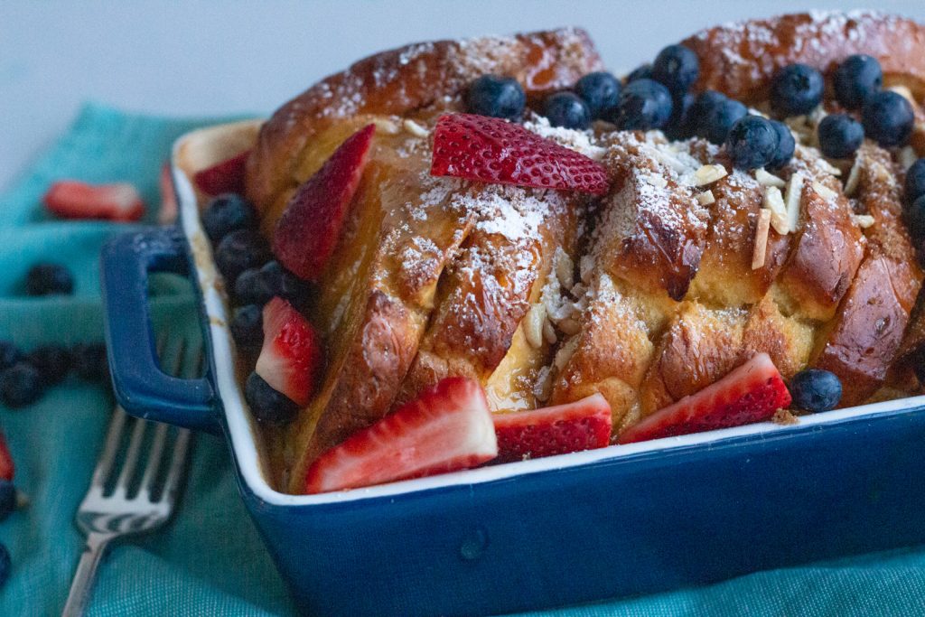 An overnight french toast bake made using brioche bread is placed in a dark blue tray with sliced almonds, strawberries and blueberries on top. The tray is kept on a blue napkin, and there is a fork on the left. 