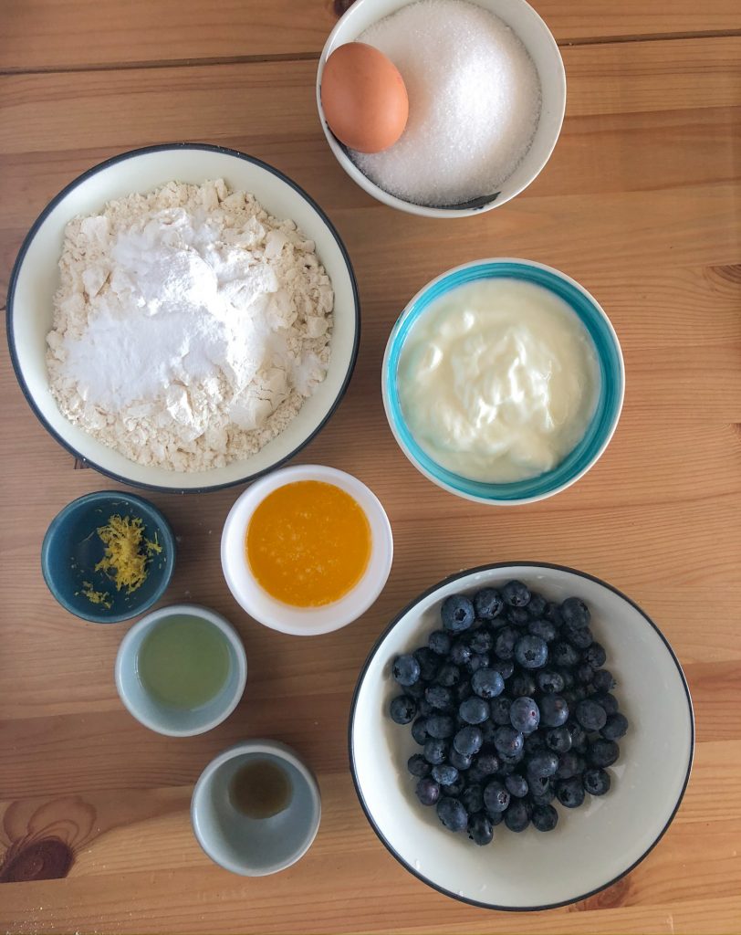 Ingredients for lemon blueberry muffins set out. Clockwise from the top is a bowl of sugar with an egg in it, then yoghurt, blueberries, vanilla essence, lemon juice, lemon zest, melted butter and flour with baking soda and salt. 