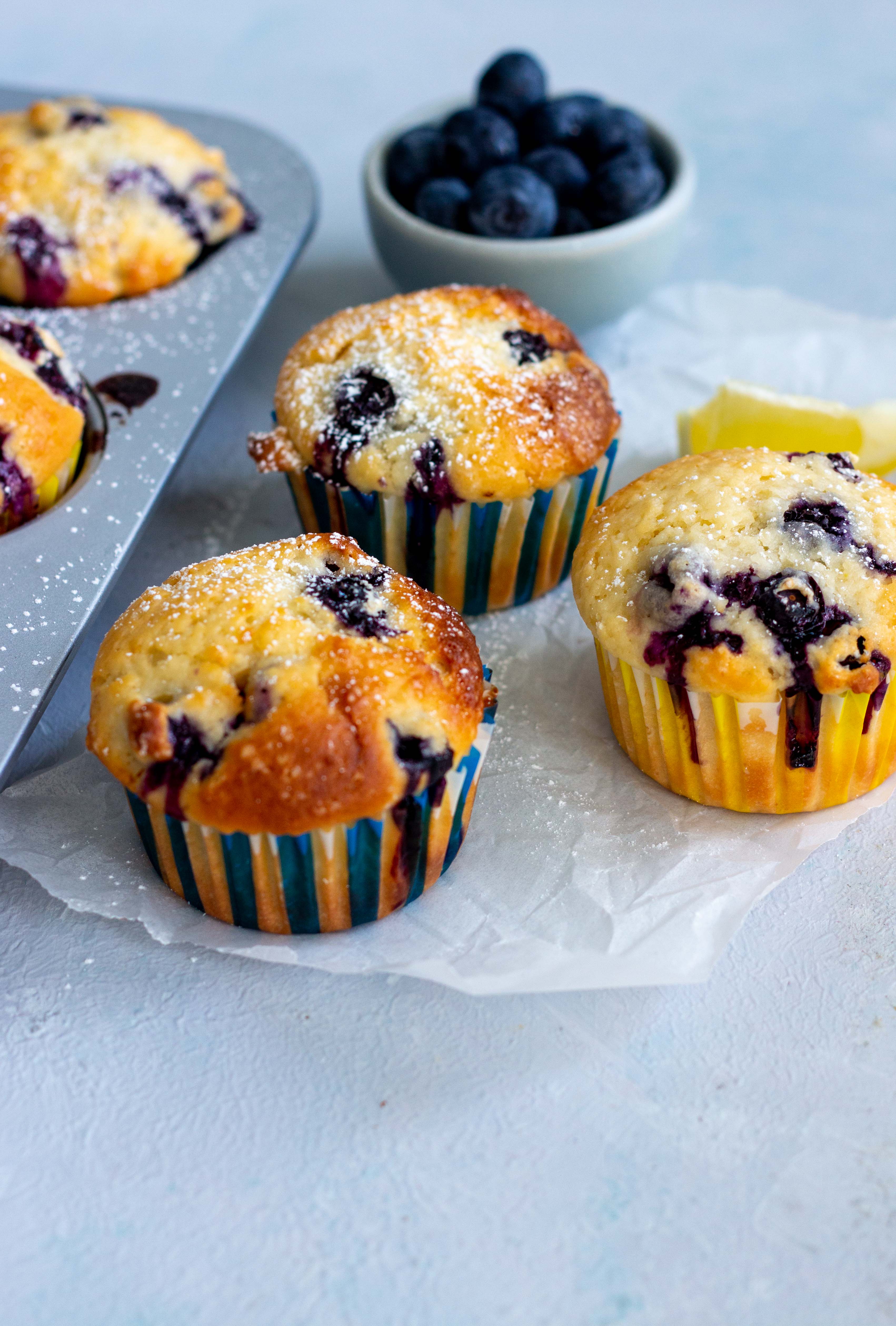 A muffin tray peeking from the left hand sided with two lemon blueberry muffins in it. Next to the tray are three lemon blueberry muffins on top of parchment paper. A mini light blue bowl with blueberries is at the back of the muffins.