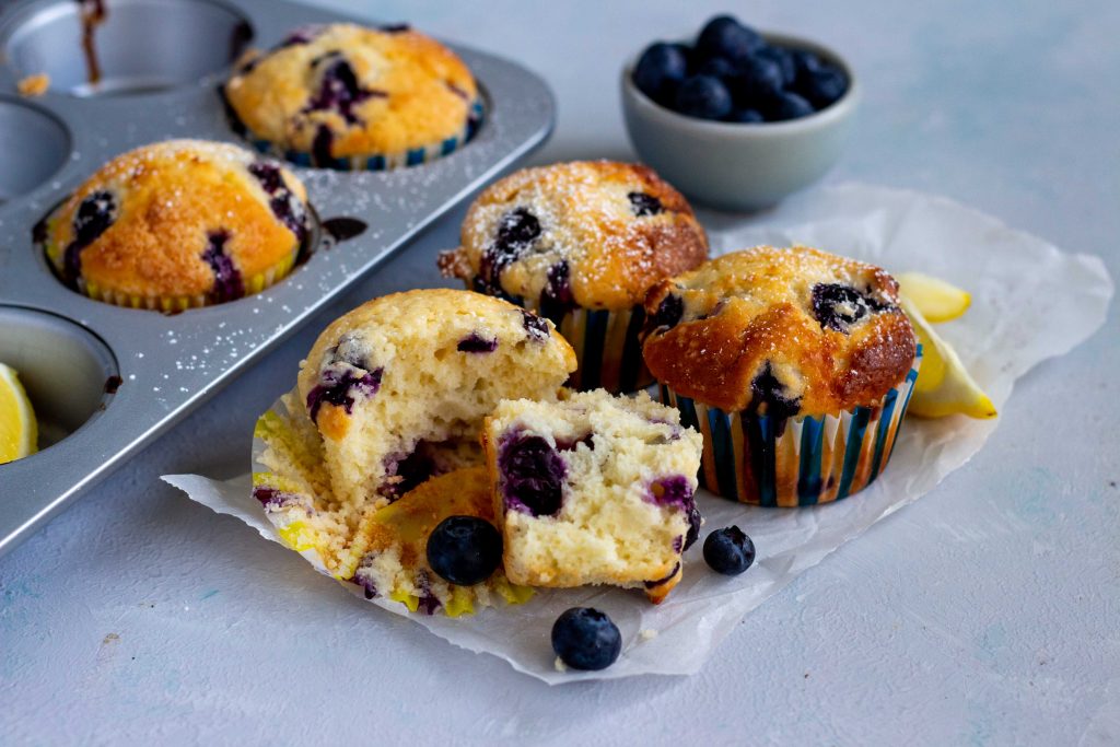 A muffin tray peeking from the left hand sided with two lemon blueberry muffins in it. Next to the tray are three lemon blueberry muffins, with the one in front half torn apart. A mini light blue bowl with blueberries is at the back of the muffins.