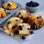 A muffin tray peeking from the left hand sided with two lemon blueberry muffins in it. Next to the tray are three lemon blueberry muffins, with the one in front half torn apart. A mini light blue bowl with blueberries is at the back of the muffins.