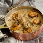 A copper coloured saucepan filled with mushroom sauce topped with five golden brown mushrooms, salt and black pepper. This copper saucepan is placed on top of a black and white striped napkin and on the top of image, the edge of a wooden spoon is peeking through.