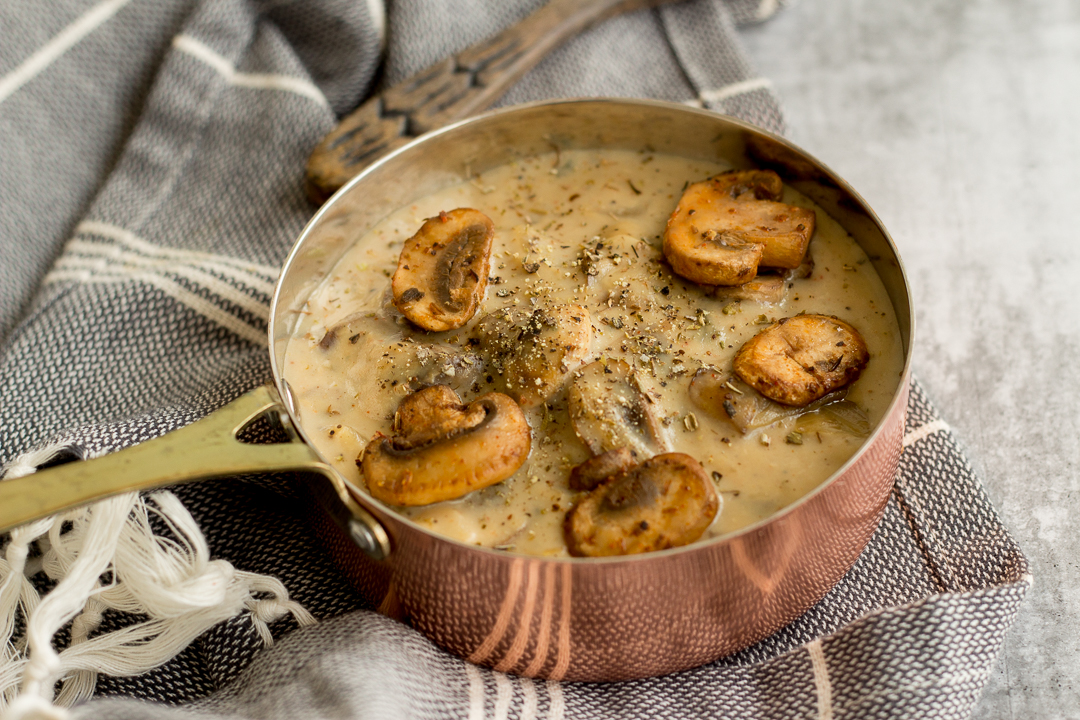 A copper coloured saucepan filled with mushroom sauce topped with five golden brown mushrooms, salt and black pepper. This copper saucepan is placed on top of a black and white striped napkin and on the top of image, the edge of a wooden spoon is peeking through.