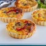 Four chicken and mushroom mini quiches with roasted capsicum on a white platter. There's a silver tong in the background.