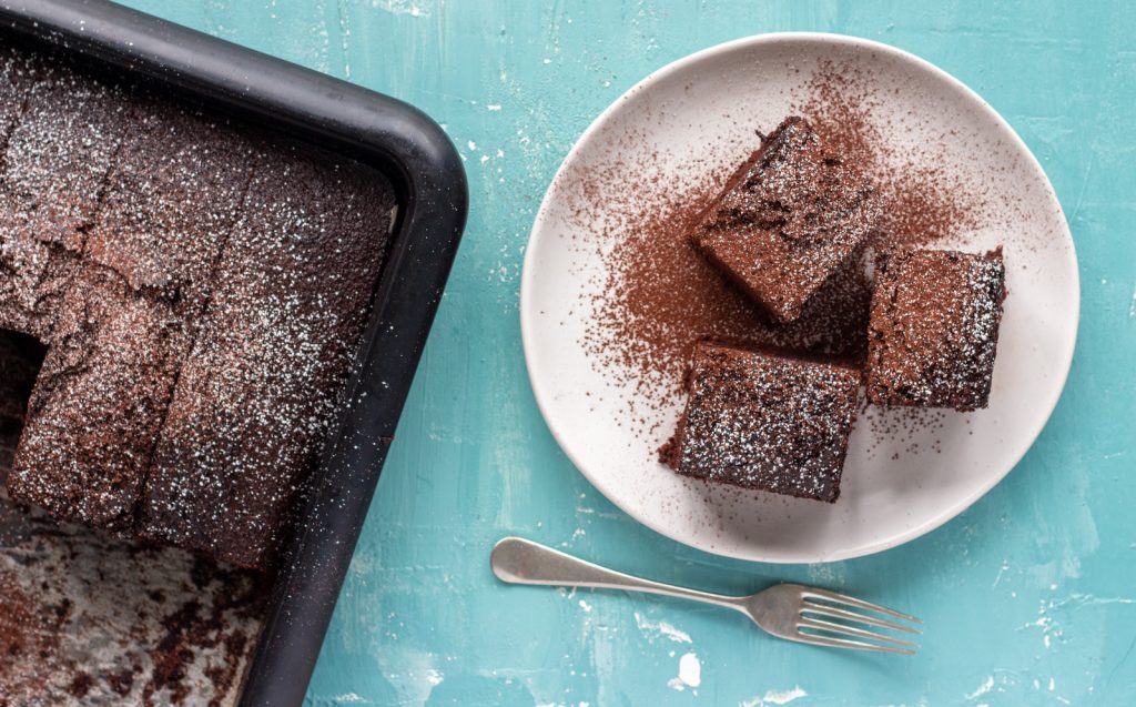 A top down shot of three slices of one bowl chocolate cake on a white plate, and dusted with icing sugar and cocoa powder. Below the plate is a silver fork. On the left of the plate is a tray with chocolate cake in it. 