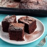 Three slices of one bowl chocolate cake on a white plate, and dusted with icing sugar and cocoa powder. On the back of the plate is a tray with chocolate cake in it.