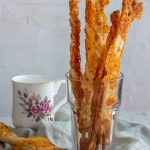 A couple of puff pastry cheese straws in a clear glass with a tea mug in the background.