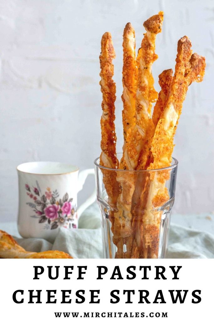 A couple of puff pastry cheese straws in a clear glass with a tea mug in the background.