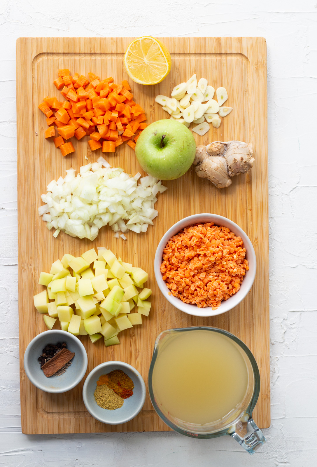 Ingredients laid out for vegetarian mulligatawny soup on a wooden chopping board. Ingredients are carrots, onions, potatoes, spices, whole spices, vegetable stock, lentils, apple, ginger, garlic and lemon. 