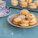 Salted caramel melting moments in a plate. There are 6 melting moments in the plate, drizzled with powdered sugar and caramel sauce. On the back is a wire rack topped with more melting moments, and a cup of tea on the left side.