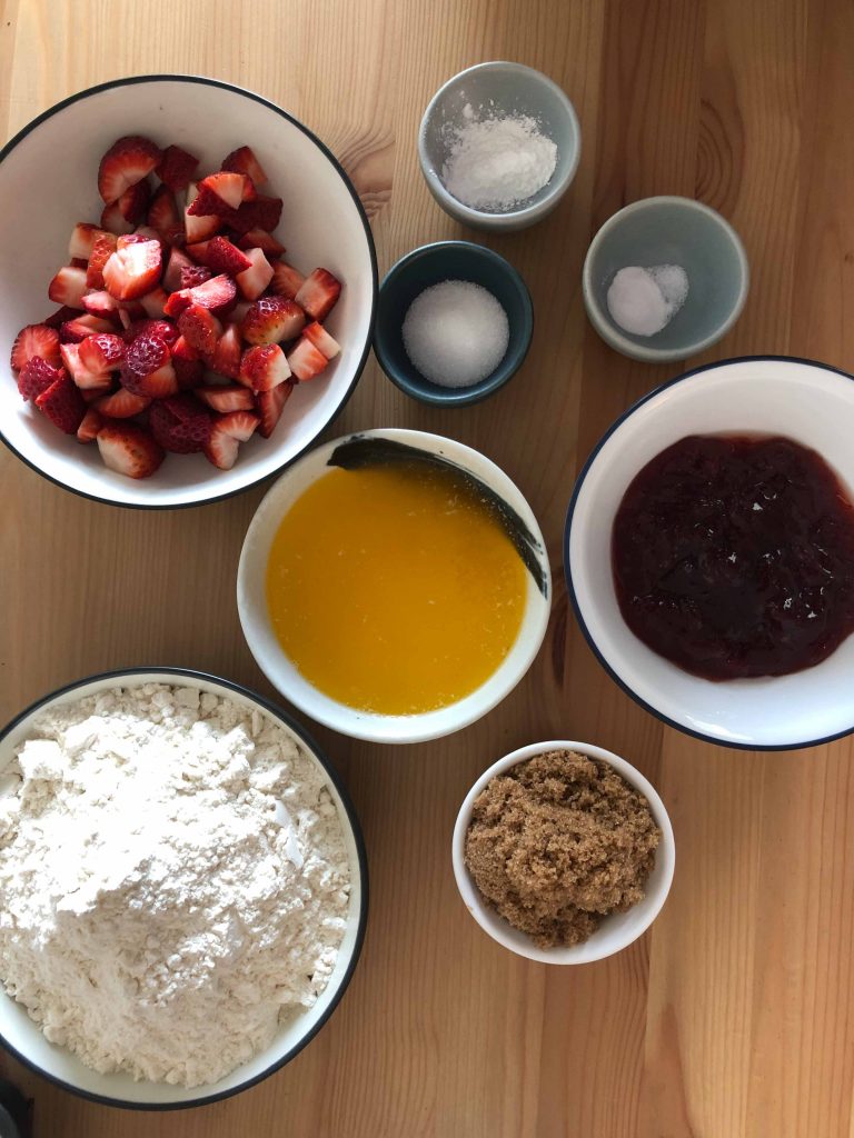 Ingredients laid out for strawberry jam bars. Ingredients are flour, brown sugar, melted butter, strawberry jam, baking soda, salt, cornflour and chopped strawberries.