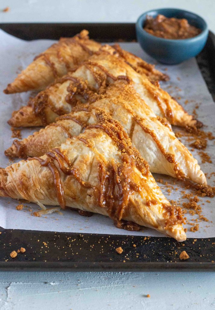 Four apple biscoff turnovers are laid out on a baking tray. They are drizzled with a lotus biscoff glaze, and topped with crushed Biscoff biscuits.