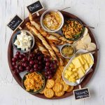 A cheese board set out for South Asians. Clockwise from the top there's cubed feta cheese, puff pastry straws, baked brie, masala peanuts, rice crackers, cubed and sliced cheddar cheese, ritz crackers, papri crisps, grapes, and mango chutney.