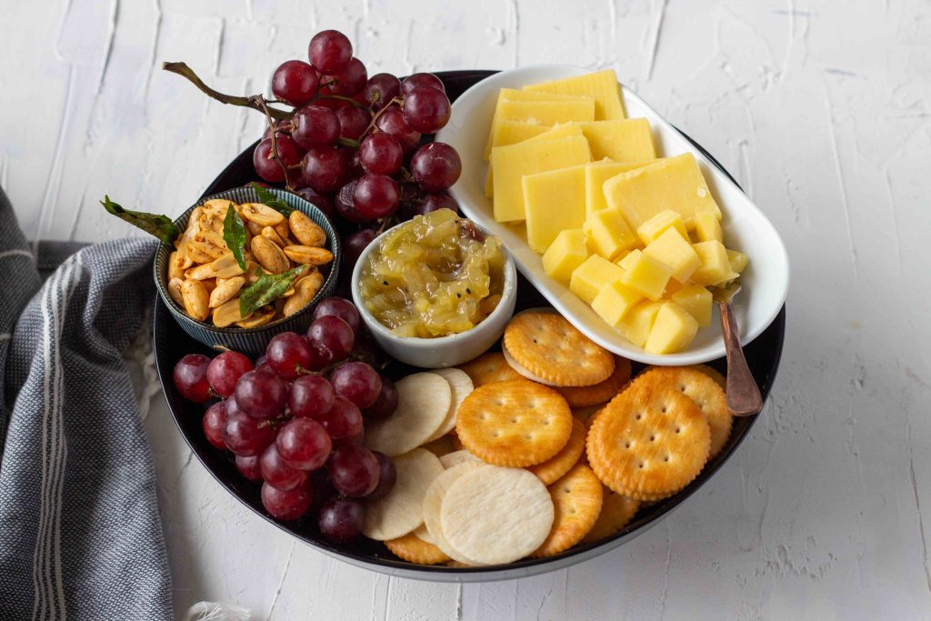 A small cheese board for around 4 people - the cheese board has grapes, masala peanuts, crackers, cubed and sliced cheddar, and mango chutney. 