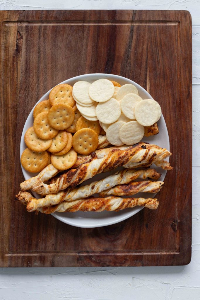 Selection of breads and crackers for a cheese board. There's Ritz crackers, rice crackers and puff pastry twists. 