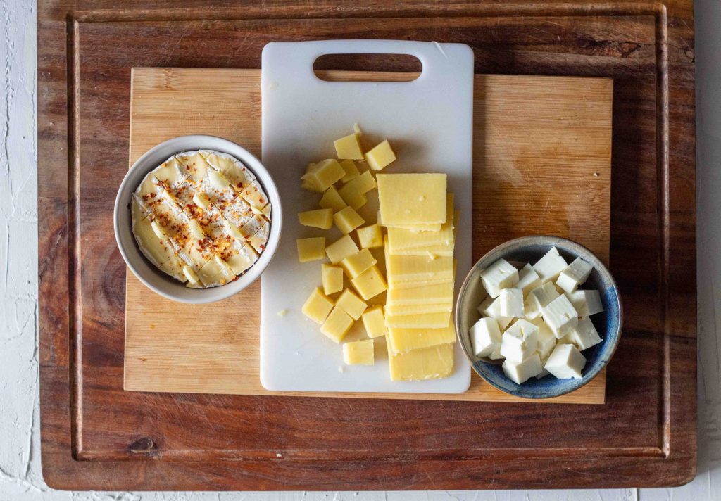 Cheese set out for a cheese board. From left to right there is brie with garlic and chilli flakes in a bowl, followed by cubed and sliced cheddar, and then cubed feta cheese.