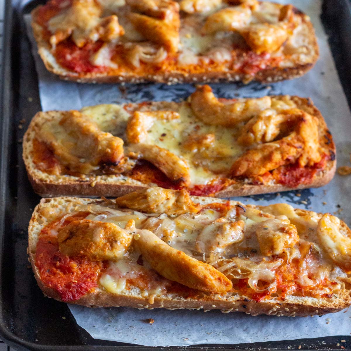 Three slices of chicken tikka bread pizza on a baking tray. The bread pizza is topped with grilled chicken flavored with chicken tikka spices, onions, tomato sauce and cheese.