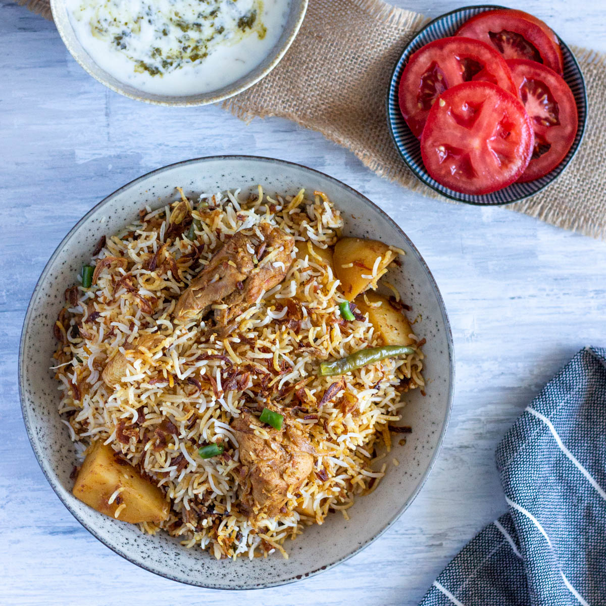 A top down image of a plate of leftover chicken biryani with potatoes. On the top right is a small plate with sliced tomatoes, and on the top left is a bowl of yoghurt raita that is half cut off from the top.