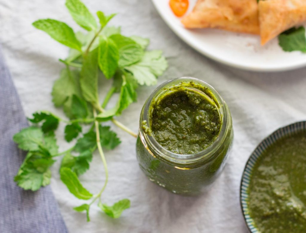 A jar of green chutney with coriander and mint next to it.