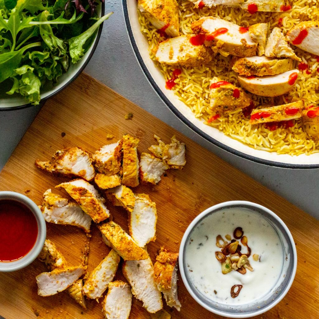 Halal cart style chicken and rice. There is grilled chicken on a chopping board with white sauce and red sauce. There is also a corner of a big pot with rice, chicken and red sauce on top peeking from the top right. On the top left corner is a bowl of salad greens, and only the edge shows. 