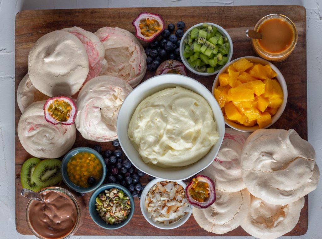 A pavlova grazing platter on a wooden chopping board. There are mini pavlovas, chopped mangoes, diced kiwifruit, passionfruit, blueberries, toasted coconut flakes, along with salted caramel sauce and chocolate ganache.