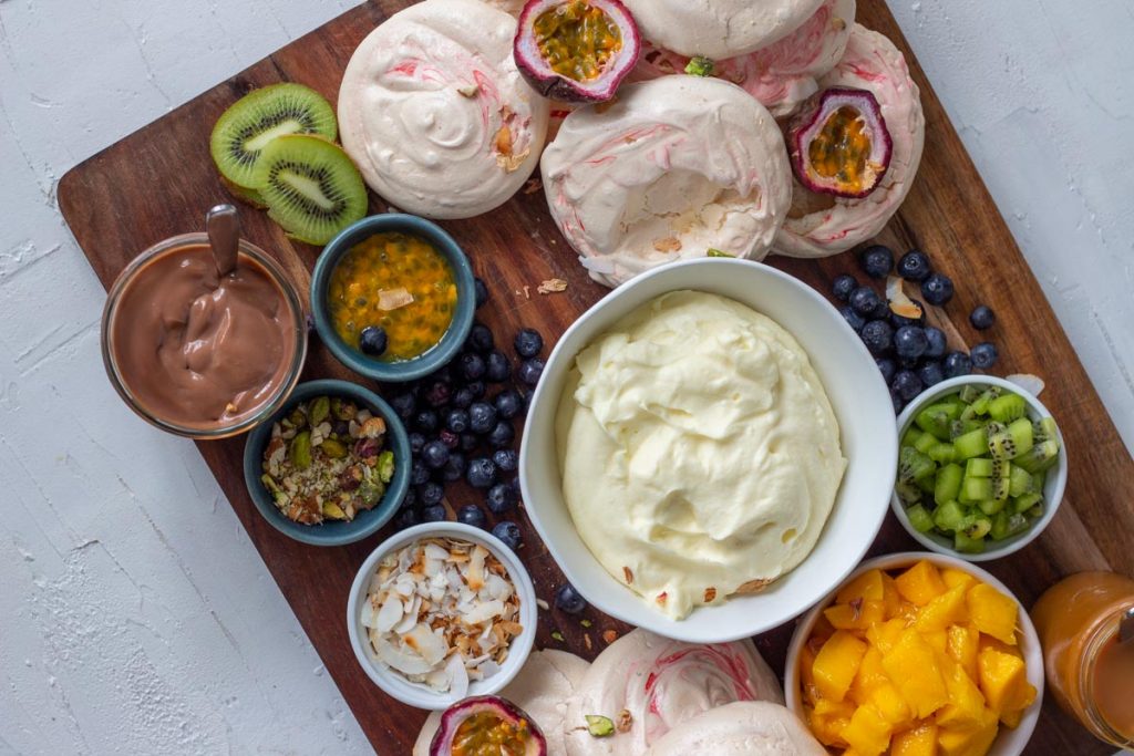 A pavlova grazing platter  at an angle on a wooden chopping board. There are mini pavlovas, chopped mangoes, diced kiwifruit, passionfruit, blueberries, toasted coconut flakes, along with salted caramel sauce and chocolate ganache.