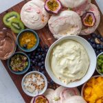 A pavlova grazing platter at an angle on a wooden chopping board. There are mini pavlovas, chopped mangoes, diced kiwifruit, passionfruit, blueberries, toasted coconut flakes, along with salted caramel sauce and chocolate ganache.