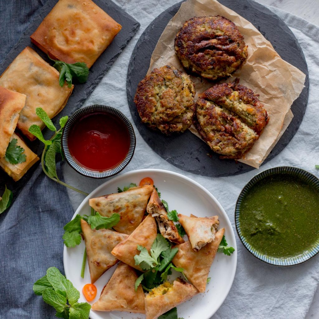 A plate of keema samosas (mince samosas), with green chutney on the side. On top of the green chutney is a black slate plate with three shami kebabs (lentil and meat kebabs) with a bowl of tomato ketchup next to it. On the top left are chicken box patties. 