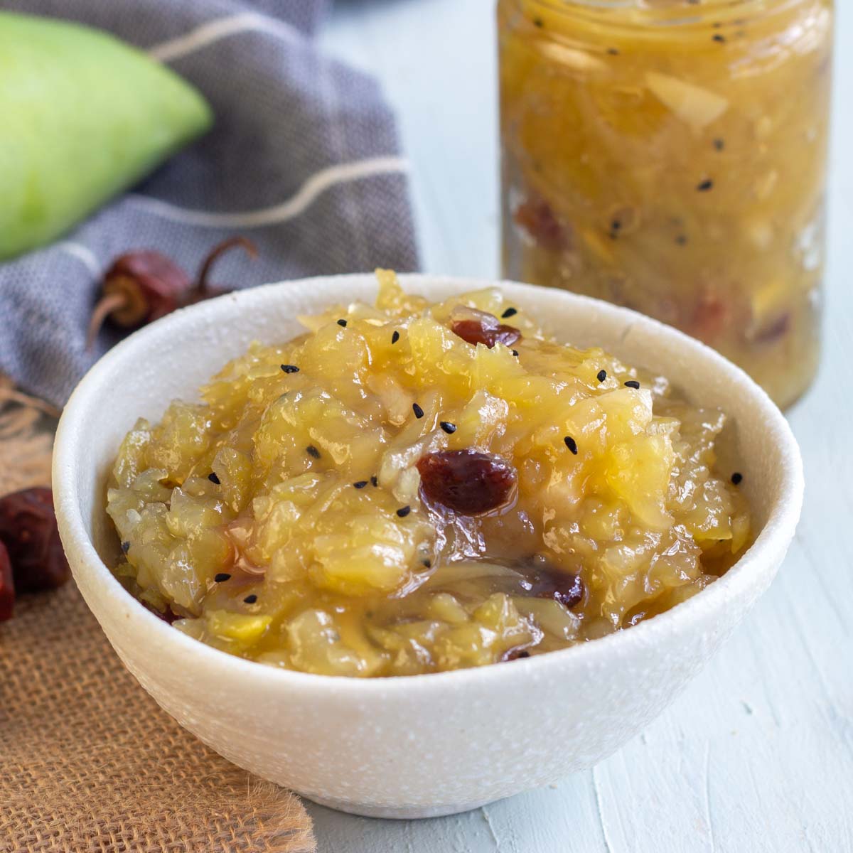 A white bowl filled with sweet mango chutney or kairi ki meethi chutney. On the back is another jar filled with mango chutney, a few whole red chilies, and a raw unripe green mango.