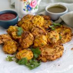 A mound of air fryer pakoras with coriander on top. Behind the air fryer pakoras is green chili sauce, and tomato ketchup.