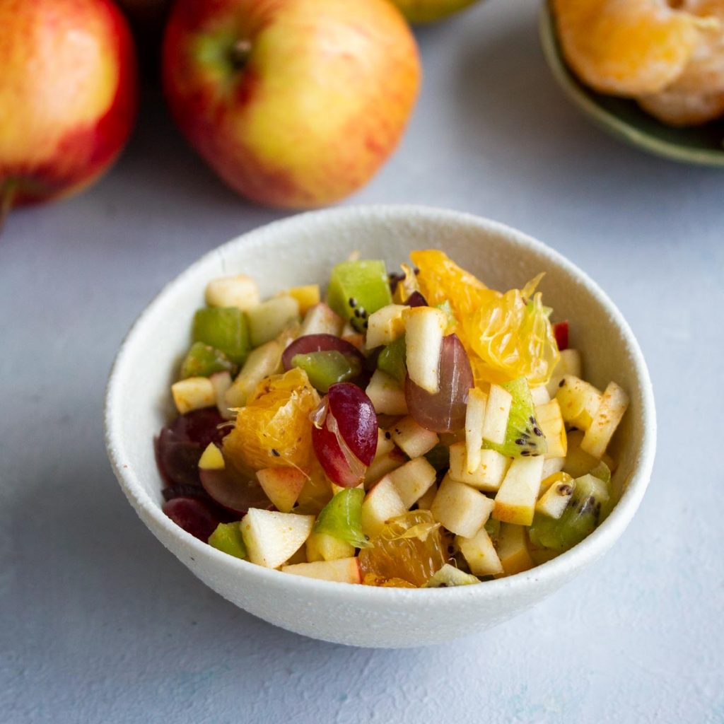 A bowl of fruit chaat containing diced apples, diced oranges, diced grapes, and diced kiwifruit in a white bowl on a white background.