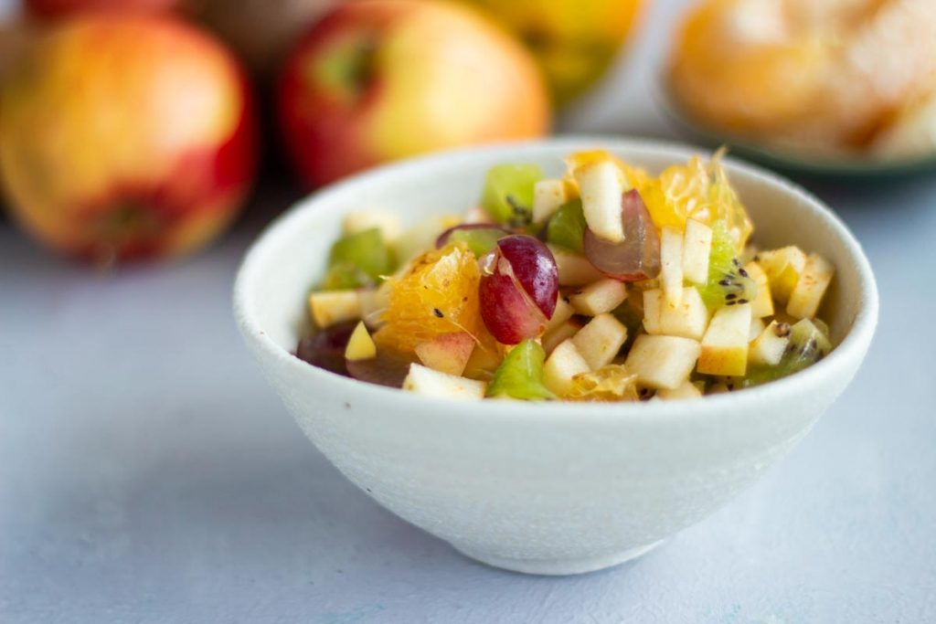 A bowl of fruit chaat containing diced apples, diced oranges, diced grapes, and diced kiwifruit in a white bowl on a white background.