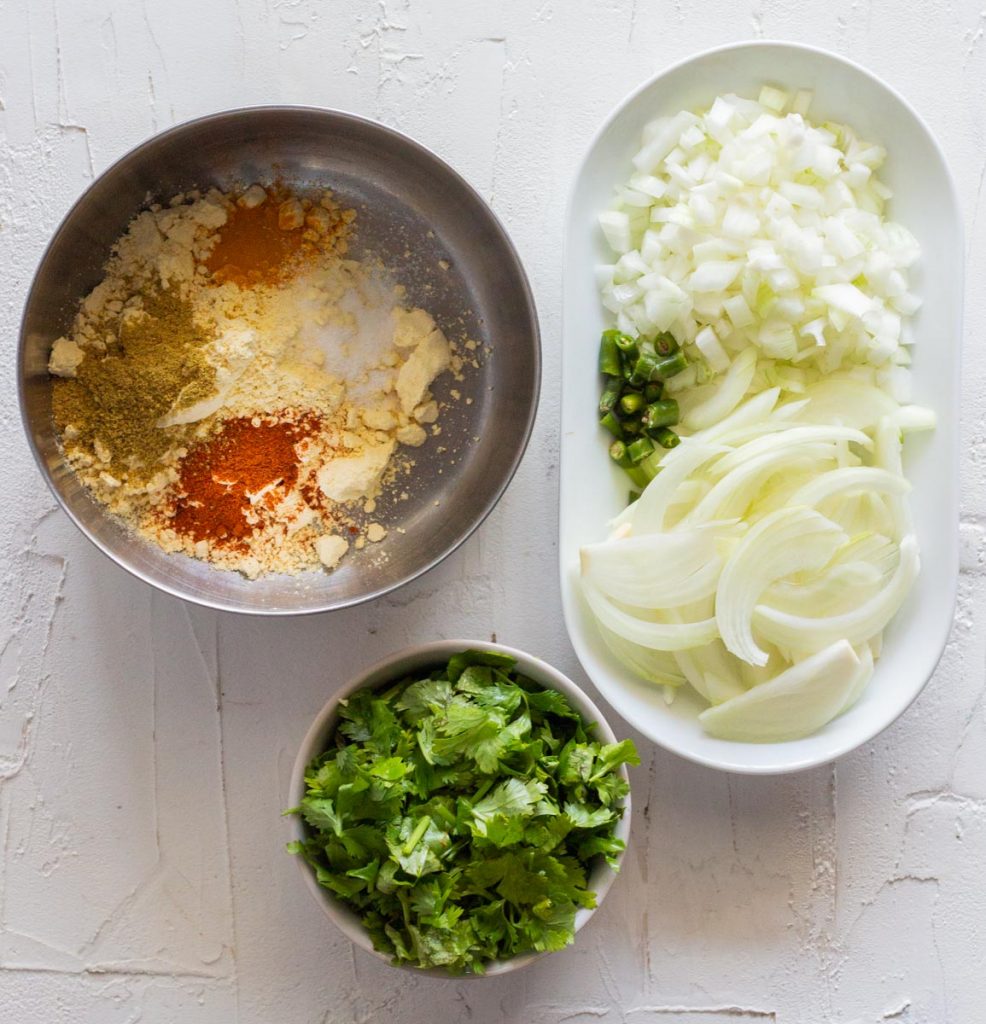 Ingredients for air fryer pakoras - chopped coriander leaves in a small bowl and a large bowl of chickpea flour / besan with red chili powder, salt, coriander powder and turmeric. There is also a plate of sliced onions, chopped onions and green chilies. 