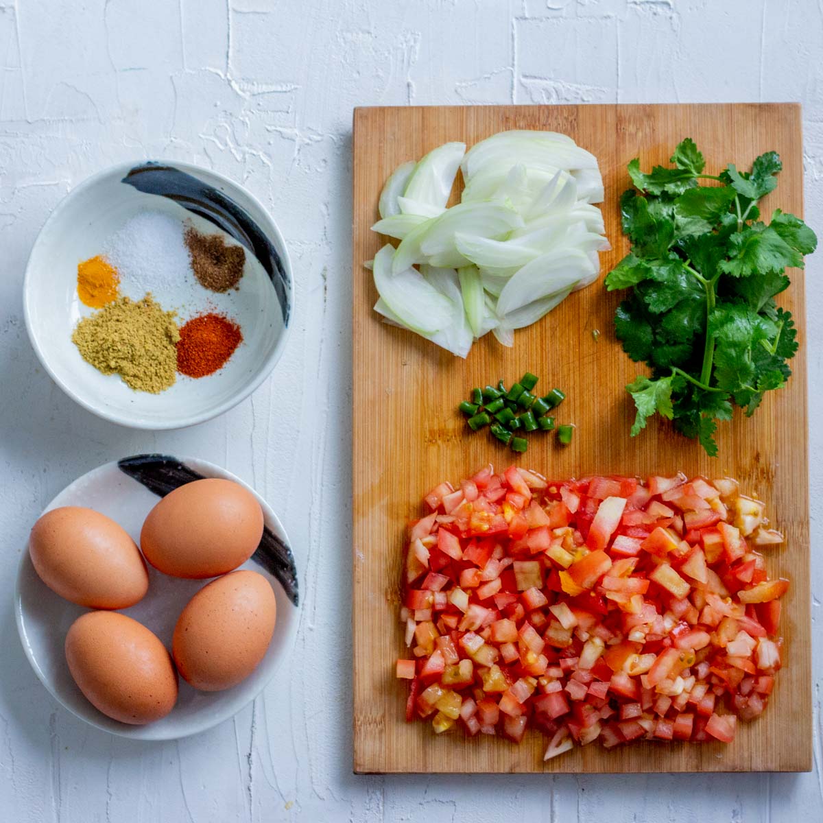 On the bottom left is a small plate with 4 eggs. Above the plate is a small bowl with coriander powder, red chili powder, salt, turmeric powder and garam masala. On the right is a chopping board with chopped tomatoes, sliced onions, green chilies and coriander leaves kept on top of it. 