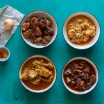 Four white bowls with blue rims. Clockwise from the top is dry chicken curry, boneless chicken curry (chicken ka salan), goat meat / mutton ka salan, and then bone in chicken ka salan.