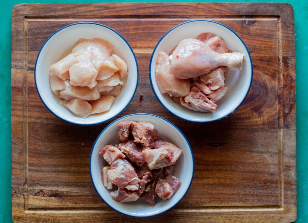 Three white bowls with blue rims. Clockwise from the top is boneless chicken, bone in chicken pieces, and then goat meat / meat.