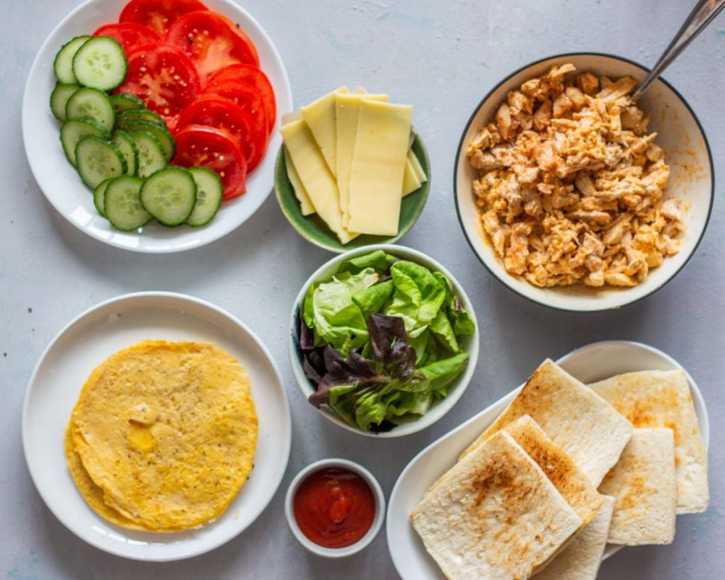 Ingredients laid out for chicken club sandwich. Clockwise from the top is sliced cucumber, and sliced tomatoes in a plate, sliced cheese in a green plate, shredded chicken with sriracha sauce, toasted bread slices, tomato sauce in a small bowl, thin omelets in a plate, and lettuce in a small bowl. 