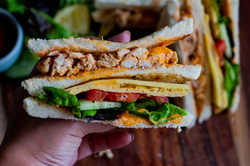 A person holding up a chicken club sandwich so that the cross section is visible. There is bread, chicken, sriracha mayonnaise, sliced cucumber, sliced tomato, cheese and lettuce.