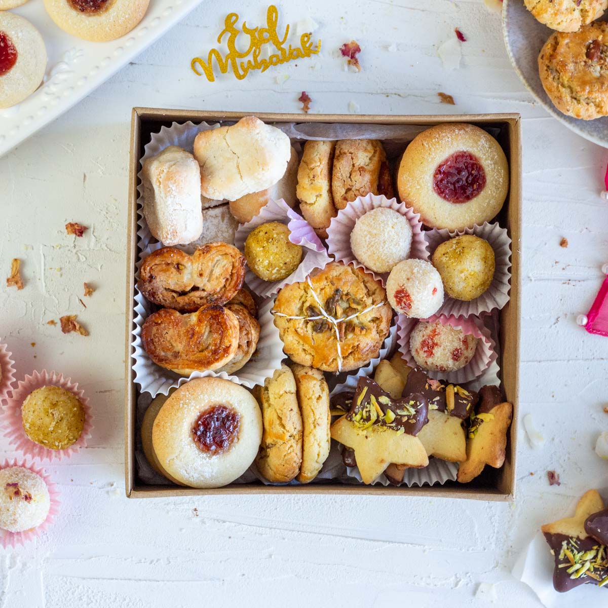 An Eid cookie box with an assortment of cookies. Clockwise from top left are Italian almond cookies, nan khatai, thumbprint jam cookies, coconut laddo, cardamom shortbread cookies with dark chocolate and pistachio, nan khatai and cinnamon palmers with nuts.