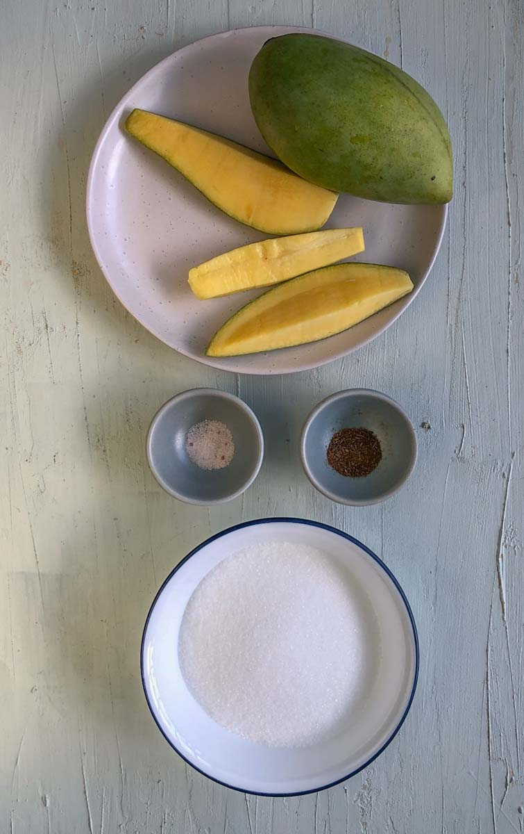 A white plate with a green mango, and three wedges of raw green mango. Below that are two mini bowls, one with roasted cumin powder, and one with black salt. On the bottom is a bowl of white sugar.
