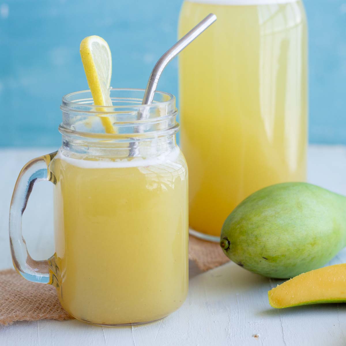 A mason jar with aam panna or green mango drink with a slice of lemon, and a stainless steel straw in it. Behind the mason jar is a bottle filled with aam panna, and a raw green mango.