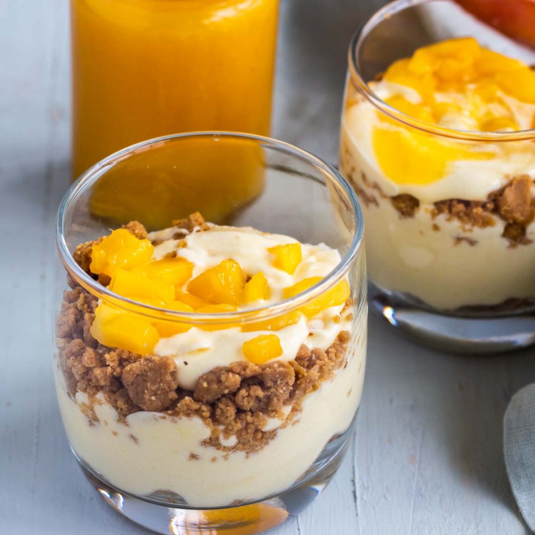 Two jars of no bake mango cheesecake. There is a biscuit crumb on the bottom, topped with a mango cream cheese filling, and chopped mangoes on top. At the back is mango puree in a glass jar.